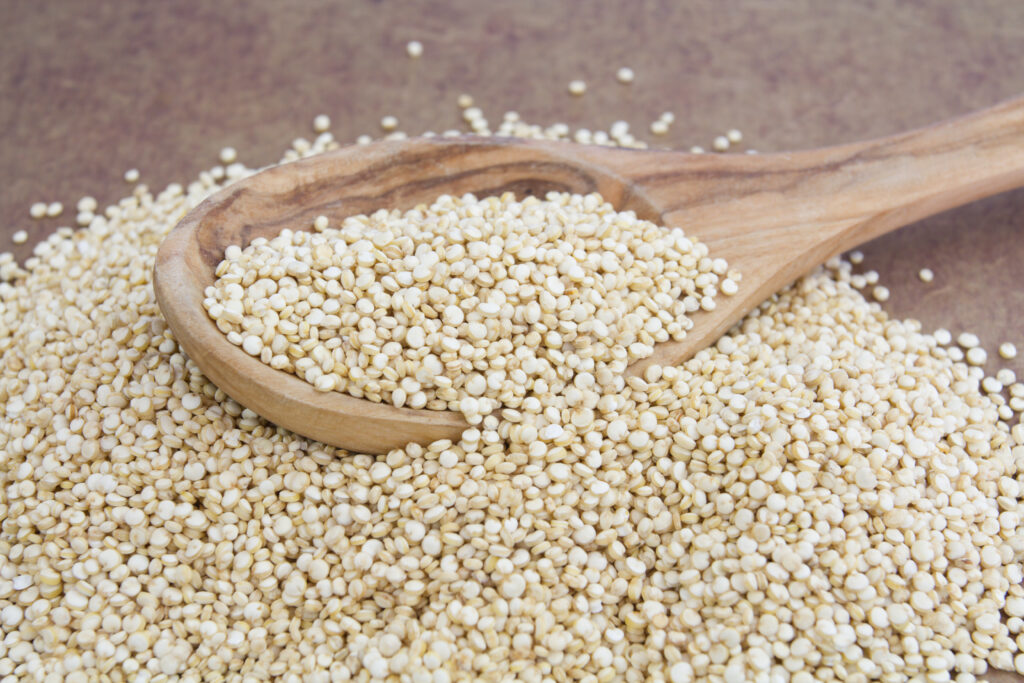 A wooden spoon overflowing with raw quinoa grains, resting on a mound of quinoa against a neutral-toned background.