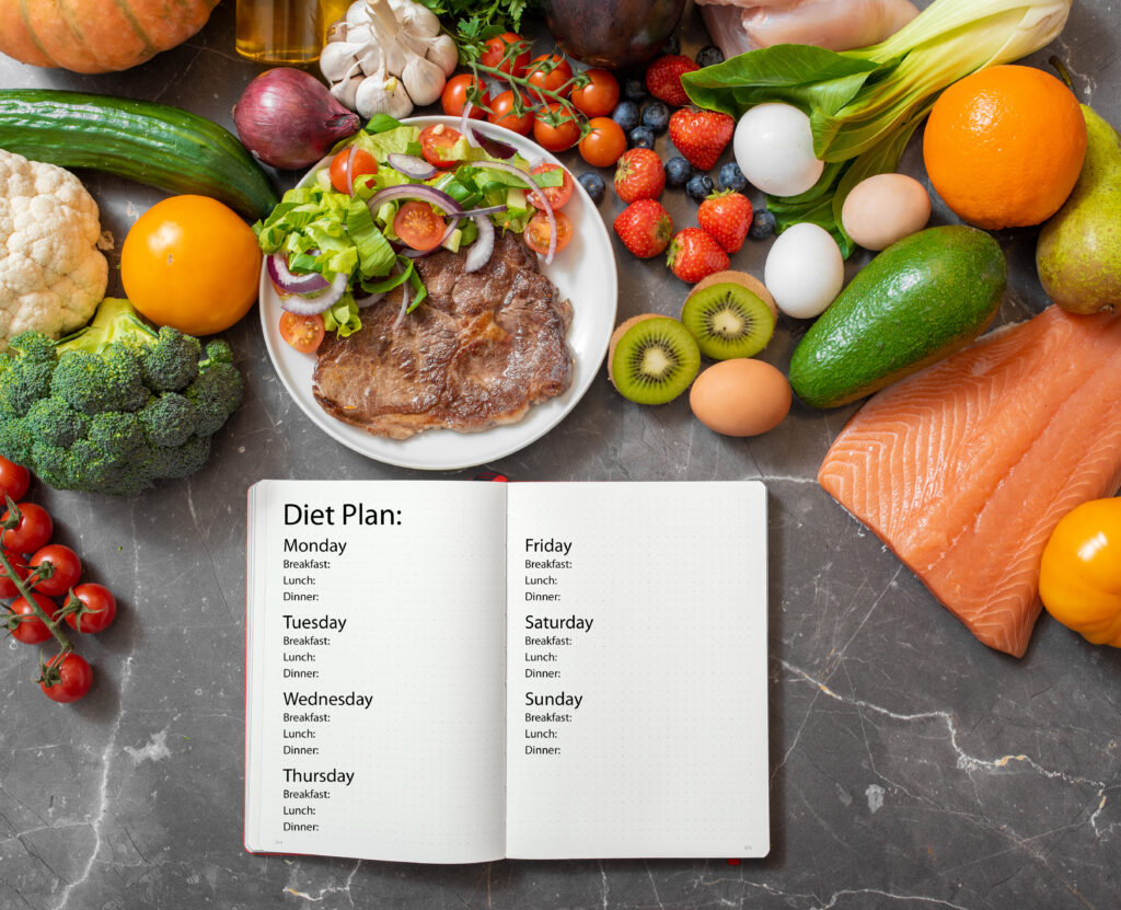 An open diet plan notebook rests on a kitchen counter surrounded by a diverse array of fresh foods including vegetables, fruits, and proteins, symbolizing the First Healing Steps for Gastritis.