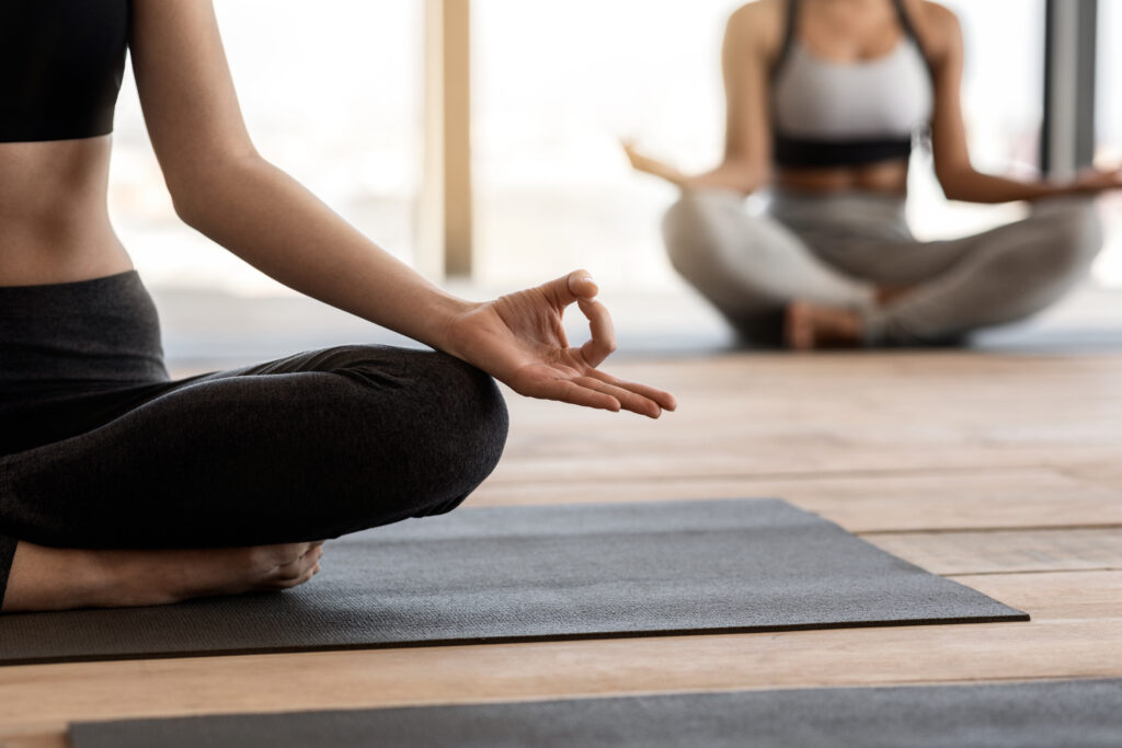 Close-up of a person in a peaceful yoga pose, hands gently resting on knees in a mudra, with another individual meditating in the background, symbolizing stress management, inner peace, and the importance of mindfulness for a positive outlook on the gastritis healing journey.