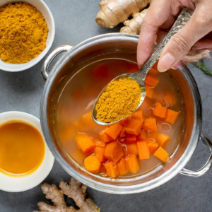 A hand adding a spoonful of vibrant ground turmeric to a pot of simmering carrots and broth, infusing the dish with its distinctive color and health-boosting properties for a flavorful carrot and ginger soup.