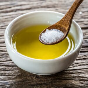 A wooden spoon sprinkling coarse sea salt into a bowl of golden olive oil, set on a textured wooden surface for a simple, homemade salad dressing.