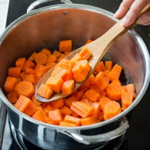 Chopped carrots being stirred in a shiny pot with a wooden spoon, the start of cooking for a nourishing carrot and ginger soup, ideal for a healthy gastritis diet.