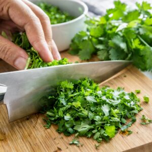 A person finely chopping fresh parsley and coriander on a wooden cutting board, preparing herbs for a flavorful dish.