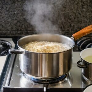 A medium saucepan on a stove with quinoa being cooked, steam rising as water boils and the heat is reduced to simmer.