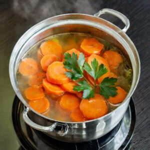 Steaming pot of sliced carrots in a clear broth, garnished with fresh parsley, simmering on the stove, ready for the next steps in a wholesome carrot and ginger soup recipe.