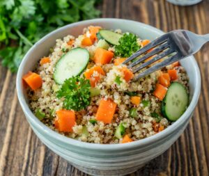 A bowl of freshly made gentle for gut quinoa salad with diced carrots, sliced cucumber, and a garnish of parsley, with a fork poised to take a bite, all set on a rustic wooden table with fresh parsley.