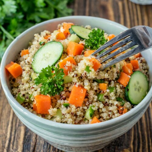 A bowl of freshly made gentle for gut quinoa salad with diced carrots, sliced cucumber, and a garnish of parsley, with a fork poised to take a bite, all set on a rustic wooden table with fresh parsley.