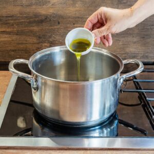 A hand pouring a tablespoon of golden olive oil into a large stainless steel pot on the stove, signifying the first step in preparing a savory carrot and ginger soup.