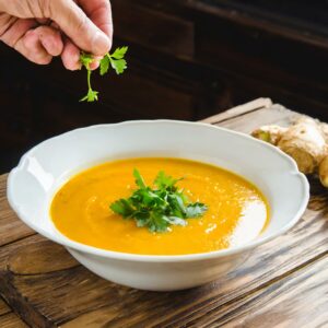 A hand garnishing a smooth, golden carrot and ginger soup with a sprig of fresh coriander, highlighting the final touch of freshness in this healthy, comforting dish.