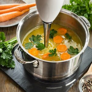 An immersion blender mixing a pot of tender carrots and fresh parsley, blending into a smooth and creamy carrot and ginger soup, rich in color and nutrition.