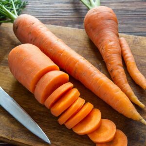 Fresh carrots being chopped on a wooden cutting board, with one carrot sliced into even rounds next to a sharp kitchen knife, ready for use in a nutritious carrot and ginger soup