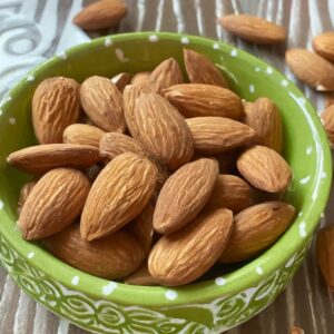 A small green bowl filled with whole, raw almonds on a wooden surface, suggesting a step in the recipe where almonds are measured before being toasted to add crunch to a dish.