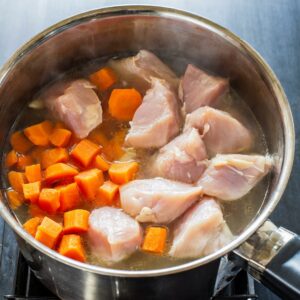 Bright orange carrot cubes added to a simmering pot with tender chicken pieces, the broth taking on a golden hue as the flavors begin to combine.