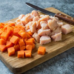 Chopped raw carrots and cubed chicken breast neatly arranged on a wooden cutting board, with a kitchen knife to the side, ready for cooking.