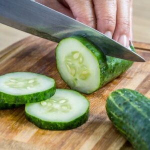 A close-up of a hand slicing a fresh, green cucumber into thick rounds on a wooden chopping board, with several slices already lying flat next to the cucumber.