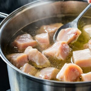 Cubed chicken pieces gently simmering in a large stainless steel pot, with a ladle stirring the clear broth as steam rises softly above.