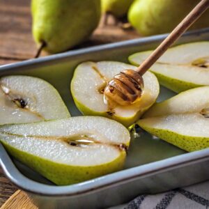 A honey dipper elegantly drizzles Manuka honey over ripe pear halves in a baking dish, accentuating the natural sweetness of the fruit with the rich, golden syrup.