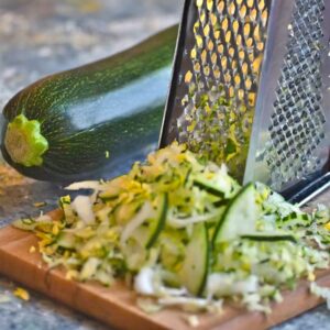 A fresh zucchini being grated on a stainless steel grater, its green skin giving way to a pile of bright, shredded flesh atop a wooden cutting board, the first step in preparing ingredients for a nutritious omelette.