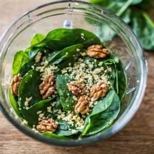 A clear glass bowl filled with vibrant fresh spinach leaves, crunchy walnuts, and a sprinkle of nutritional yeast, ready to be transformed into a nutrient-rich walnut pesto.