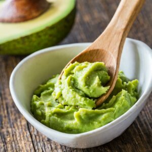 Creamy mashed avocado in a white bowl with a wooden spoon, ready to be used on Toast.