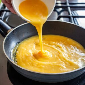 A cascade of whisked golden eggs flows smoothly from a bowl into a sizzling skillet, ready to be cooked into a soft, fluffy Gastritis-Friendly Spinach and Fennel Omelette over a gentle flame.