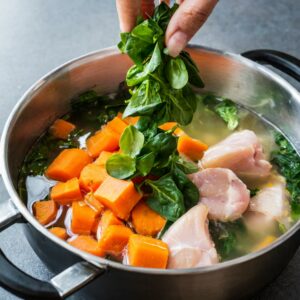 A hand adding a generous handful of fresh, vibrant spinach to a pot where cubed chicken and chopped carrots are simmering, the greens ready to wilt into the warm, nourishing broth.