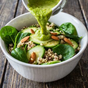 A vibrant bowl of quinoa and fresh spinach salad is being dressed with a thick, green avocado sauce poured from above, with slices of cucumber and whole almonds adding texture and nutrition to the dish.