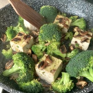 Slightly golden tofu cubes combined with fresh, green broccoli and savory mushrooms being stir-fried together, creating a colorful and appetizing mix in a skillet.