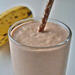 The image is of a Creamy Carob Smoothie in a glass, with a ripe banana in the background, indicating a rich and sweet beverage.