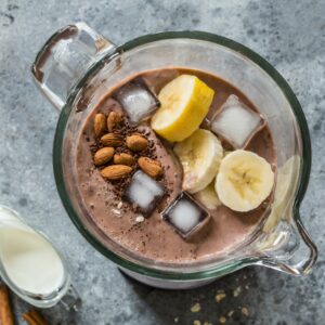 A pre-blended smoothie mixture with banana slices, almonds, carob powder, and ice cubes in a blender, ready for blending to create a creamy carob smoothie