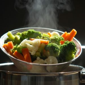 Steam rises from a colander full of vibrant, freshly boiled vegetables including broccoli, carrots, and cauliflower, held over a pot by a hand, showcasing a healthy, gastritis-friendly cooking method.