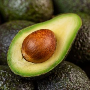 A ripe avocado cut in half revealing its creamy flesh and pit, surrounded by a group of whole avocados, epitomizing the freshness and nutritional value of raw ingredients in a gastritis-friendly diet.