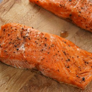 Two succulent salmon fillets seasoned with cracked black seeds, resting on a rustic wooden cutting board after being baked, exemplifying a healthy and flavorful option for a gastritis-friendly diet.